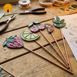 May include: A collection of stained glass fruit and leaf garden stakes. The stakes feature a red strawberry, a yellow lemon slice, red cherries, an orange slice, and green leaves. The stakes are on copper rods, and the background includes tools and materials.