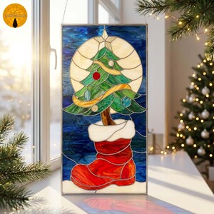 May include: A rectangular stained glass panel featuring a green Christmas tree with colorful ornaments inside a red stocking. The tree is set against a circular cream-colored background and a deep blue sky, all framed in metal with a hanging chain.