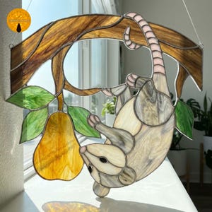 Op de afbeelding: Een glas-in-lood suncatcher met een opossum die met zijn staart aan een bruine tak hangt. De opossum is lichtgrijs en wit met roze accenten. Een felgele peer met groene bladeren hangt aan de tak vlakbij de kop van de opossum.