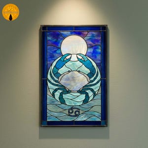 May include: Stained glass artwork featuring a blue crab design, set against a backdrop of blue and white hues. The artwork includes a white sun and the Cancer zodiac symbol. The piece is framed with a dark border.