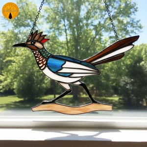 May include: Stained glass roadrunner bird suncatcher with a brown, white, blue, and red color scheme. The bird is perched on a light brown branch and hangs from a chain. The suncatcher is displayed in front of a window.