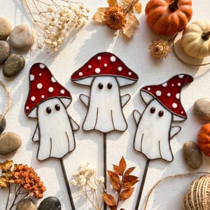 May include: Three stained glass garden stakes featuring white ghost-shaped bodies with red mushroom caps adorned with white polka dots. Each ghost has two small black eyes and is attached to a thin metal rod, creating a decorative set.