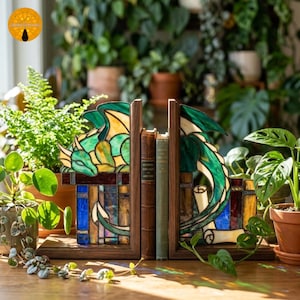 Può includere: Una coppia di fermalibri in vetro colorato con un design a drago verde con accenti gialli e blu, inseriti in cornici di legno scuro. Il drago è raffigurato in posizione di riposo su un lato e avvolto sull'altro, mentre tiene un rotolo.