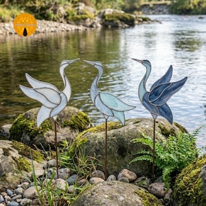 May include: Three stained glass heron garden stakes in shades of white, blue, and teal stand on a mossy rock near a body of water. The heron sculptures have outstretched wings and are mounted on metal rods for easy placement in a garden.