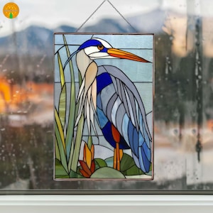 May include: Stained glass panel featuring a great blue heron. The bird is depicted with blue, gray, and white feathers, an orange beak, and yellow legs. The background includes reeds and a light blue sky. The panel is framed in a dark metal.