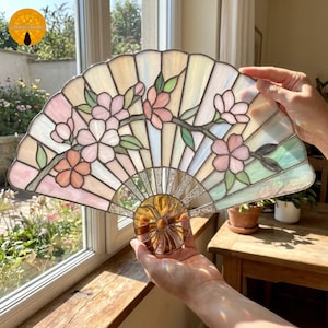 May include: A fan-shaped stained glass panel featuring iridescent white glass segments and a floral motif with pink and peach-coloured blossoms and green leaves. The piece is constructed with dark metal soldering lines and a circular amber glass centre.