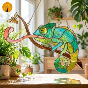 May include: A stained glass suncatcher featuring a green and yellow chameleon perched on a brown branch with its tongue extended towards a small yellow insect. The piece is constructed with metal solder lines and hangs from a silver chain.