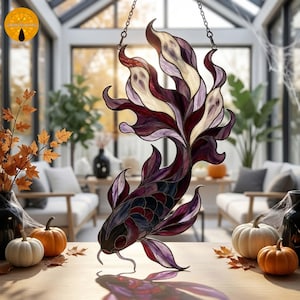 May include: A stained glass suncatcher shaped like a koi fish with deep purple, burgundy, and cream-colored glass panels. The fish features flowing, elongated fins and is suspended by a metal chain, casting a colorful shadow on the surface below.
