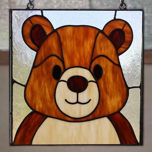 May include: Stained glass panel featuring a friendly bear face. The bear has brown fur, black eyes, and a cream-colored muzzle. The panel is square, with a black border and hanging chain, suitable for home decor.