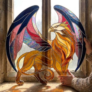 May include: A stained glass griffin, featuring a golden body, white head, and wings in blue, pink, and red. The artwork is displayed in front of a window, highlighting the detailed design and vibrant colours of the mythical creature.