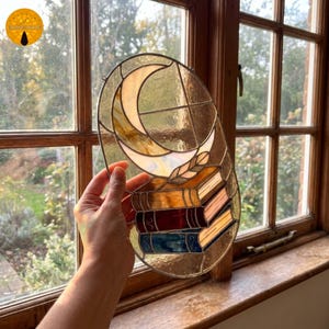 Puede incluir: Un atrapasoles ovalado de vidrieras que muestra una luna creciente sobre una pila de tres libros. La luna es de color crema, mientras que los libros están representados en tonos ámbar, rojo oscuro y azul con detalles de vidrio transparente.
