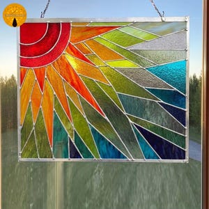 May include: Stained glass panel depicting a vibrant sunrise. The artwork features a red sun, radiating orange and yellow rays, with a background of green, blue, and gray geometric shapes. The panel is framed in silver and hangs from a chain.