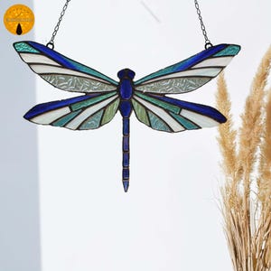 May include: A stained glass dragonfly suncatcher with a chain for hanging. The dragonfly features blue, white, and green glass panels. The wings have a striped pattern. The body and head are a solid blue colour. The suncatcher is a decorative item.