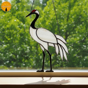 May include: Stained glass crane with a red head, black neck and legs, and white body. The bird is hanging from a chain in front of a green, blurred background. The crane is a decorative item.