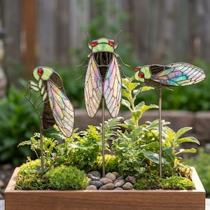 May include: Three stained glass cicada garden stakes featuring iridescent wings, light green bodies, and small red eyes. Each insect is mounted on a thin metal rod, designed to be placed upright in soil or a planter for decorative outdoor display.