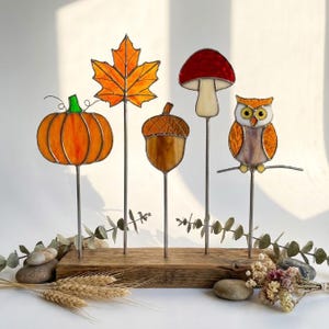 Può includere: Un set di cinque ornamenti in vetro colorato su aste metalliche inserite in una base rettangolare in legno. Gli ornamenti raffigurano una zucca arancione, una foglia d'acero gialla, una ghianda marrone, un fungo rosso e un gufo arancione.