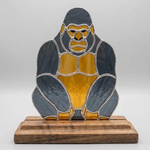 May include: Stained glass gorilla sculpture in shades of gray and yellow, with a silver-colored metal outline. The gorilla is seated on a wooden base. The sculpture is a decorative item.
