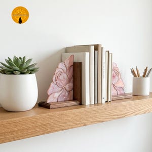 Könnte beinhalten: Dekorative Buchstützen mit floralem Design. Die Buchstützen zeigen eine rosa-weiße Glasmalerei-Blume auf einem Holzsockel. Bücher stehen zwischen den Buchstützen auf einem Holzregal. Links steht ein weißer Topf mit einer Sukkulente.