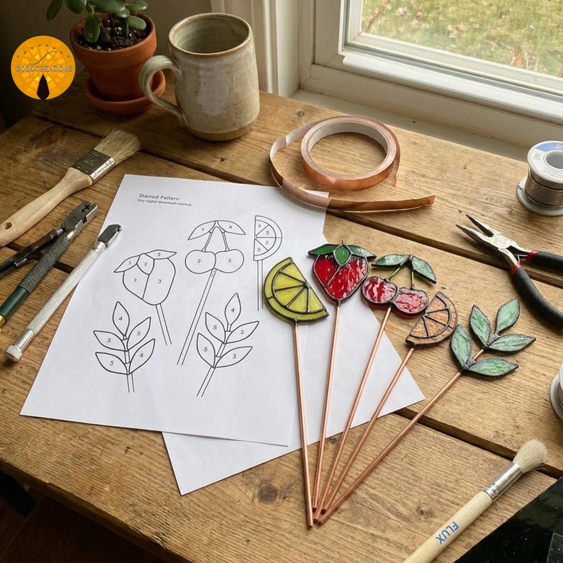 May include: A collection of stained glass fruit and leaf garden stakes, including a strawberry, lemon, cherries, and orange slice. A pattern for the stakes is on a white sheet of paper. Crafting tools and supplies are also visible.