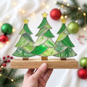 以下が含まれることがあります： 上部に黄色い星が付いた3本の緑色のクリスマスツリーをあしらったステンドグラスの装飾品で、明るい色の長方形の木製ベースに固定されています。ツリーはさまざまな色合いの緑色のガラスで構成されており、金属製のハンダ付けの線が見えます。