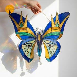Può includere: Farfalla in vetro colorato con tonalità blu, gialle e verdi. Il design incorpora motivi di occhi egizi e simboli ankh. Ha una catena dorata per appenderla e una perla di cristallo pendente.