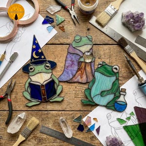 May include: Three stained glass frog figures on a wooden surface. One frog wears a blue wizard hat and reads a book, one wears a purple cloak and holds a crystal ball, and one sits beside a blue potion bottle. Tools and glass shards surround them.
