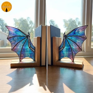 Puede incluir: Sujetalibros con forma de alas de dragón con alas de vidrieras en tonos azules, morados y rosas. Las alas están unidas a bases de madera. Los sujetalibros sostienen dos libros con cubiertas azules.