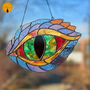 May include: A colourful stained glass dragon eye suncatcher. The eye has a black pupil and is surrounded by green, yellow, red, orange, blue, and purple glass pieces, framed by a dark metal border. It hangs from a chain, suitable for window or wall decoration.