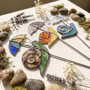 May include: Three stained glass snail ornaments on metal stakes. Each snail features a unique shell colour pattern, including brown, orange, and blue, paired with a matching umbrella. The glass pieces are joined with dark metal solder lines.