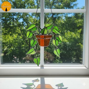 May include: Stained glass hanging plant with green leaves and a brown pot with an orange stripe. The plant is suspended by a black chain in front of a window with a view of trees. The glass has a glossy finish.