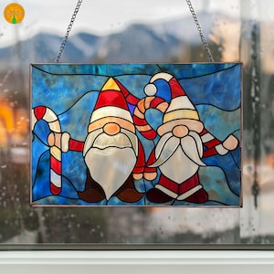 Puede incluir: Panel de vidrieras con dos gnomos navideños con barbas blancas, gorros rojos y bastones de caramelo. Los gnomos están sobre un fondo azul, y el panel está enmarcado con un borde de cobre. Una cadena está unida para colgar.