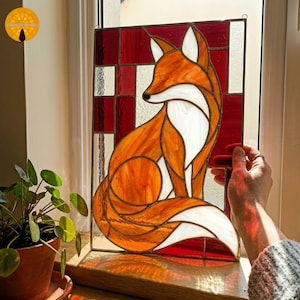 May include: Stained glass panel featuring a stylized fox in shades of orange, white, and brown. The fox is set against a red and white geometric background. The artwork is framed with a dark metal border, suitable for window or wall decor.