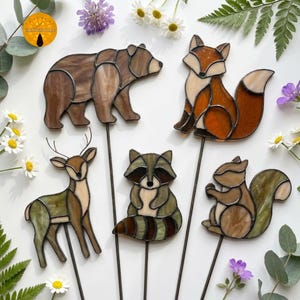 May include: A selection of stained glass animal garden stakes. The set features a bear, fox, deer, raccoon, and squirrel. Each animal is made with brown, orange, and green glass, and attached to a metal stake for display in a garden or flower bed.