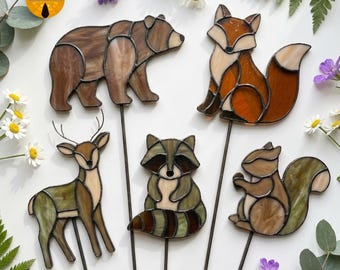 May include: A collection of stained glass animal garden stakes. The set includes a bear, fox, deer, raccoon, and squirrel. Each animal is crafted with brown, orange, and green glass, and mounted on a metal stake for easy placement in a garden or flower bed.