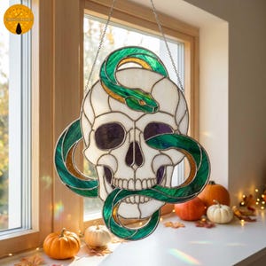 Op de afbeelding: Een glas-in-lood suncatcher met een witte schedel verstrengeld met een groene en gouden slang. Het stuk is geconstrueerd met donkere metalen soldeerlijnen en hangt aan een dunne zilveren ketting, met doorschijnende glazen segmenten.