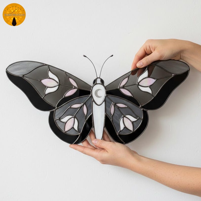 May include: A stained glass butterfly with gray, black, white, and pink details. The butterfly has a crescent moon on its body and floral patterns on its wings. The wings are a mix of gray and black, with pink and white floral accents.