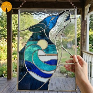 May include: Stained glass panel featuring a howling wolf silhouette against a backdrop of ocean waves and a yellow sun. The artwork uses shades of blue, teal, and white, with a clear glass border. The panel is designed to hang, with a chain at the top.