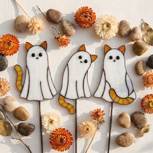 May include: Three stained glass cat figures dressed as ghosts, each featuring white bodies, orange-tipped ears, and orange striped tails. The cats are mounted on thin metal stakes and are surrounded by scattered smooth brown stones and dried orange flowers.