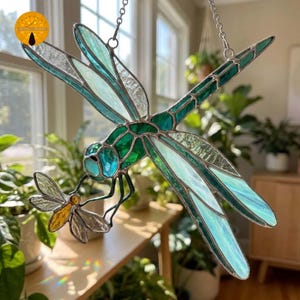 May include: A stained glass dragonfly suncatcher featuring translucent teal, green, and clear textured glass panels held together by metal solder lines. The dragonfly holds a small, yellow-bodied insect with clear wings, suspended by a silver chain.
