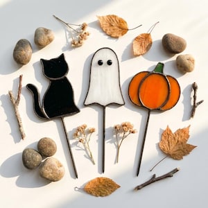 May include: Three stained glass decorative stakes featuring a black cat, a white ghost with two black eyes, and an orange pumpkin with a green stem. Each figure is mounted on a thin metal rod, arranged with scattered brown leaves and stones.