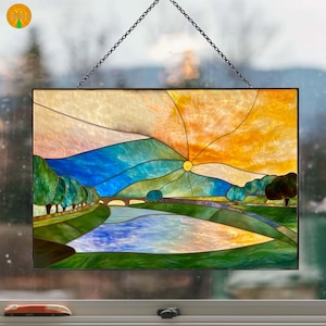May include: Stained glass panel depicting a landscape scene with a river, bridge, and trees. The sky transitions from orange to yellow, with a sunburst design. The panel is framed and hangs from a chain.