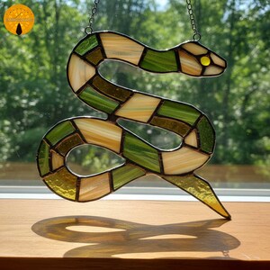 Könnte beinhalten: Bunter Glas-Sonnfänger in Schlangenform, in Grün-, Gelb- und Brauntönen. Die Schlange ist gewunden, mit einem gelben Auge. Er hängt vor einem Fenster und wirft einen Schatten auf die Holzoberfläche darunter.