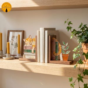 Puede incluir: Un par de sujetalibros de madera en forma de L con inserciones de vidrieras. Una pieza representa un gato naranja durmiendo sobre una pila de libros, mientras que la otra muestra una cola de gato junto a una planta en maceta y libros en tonos verdes y marrones.