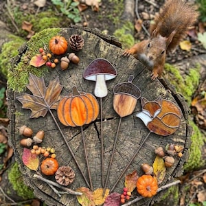 May include: Four stained glass garden stakes featuring a maple leaf, a pumpkin, a mushroom, and a fox, arranged on a tree stump with a squirrel nearby. The pieces are crafted with orange, brown, and white glass, held together by dark metal solder lines.