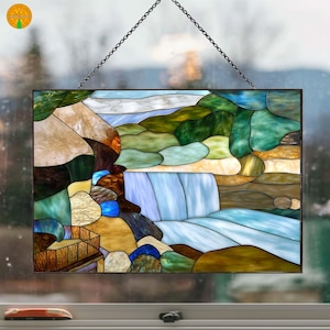 Puede incluir: Panel de vidrieras que representa una escena de cascada. La obra de arte presenta una cascada de agua azul y blanca, rodeada de rocas y follaje verdes, marrones y beiges. El panel es rectangular y cuelga de una cadena.