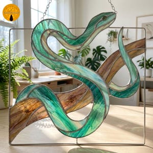 May include: A stained glass suncatcher featuring a coiled snake with translucent green and brown glass panels held together by metal solder lines. The snake is positioned within a square metal frame and hangs from a silver chain for display.