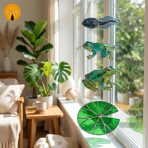 May include: A stained glass window hanging featuring a vertical chain of four pieces: a blue tadpole, a small green frog, a larger green frog, and a circular green lily pad. The pieces are connected by silver metal chains and hooks.