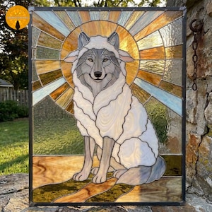May include: A rectangular stained glass panel featuring a grey wolf wearing a white sheepskin cloak. The wolf is centred against a circular amber and gold sunburst background. The piece is framed in dark metal with visible leaded glass seams.