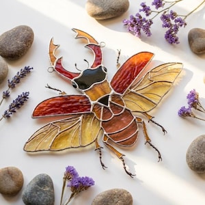 May include: A stained glass stag beetle suncatcher featuring vibrant red, orange, and yellow glass panels held together by dark metal solder lines. The insect design includes prominent mandibles, segmented wings, and thin wire legs.