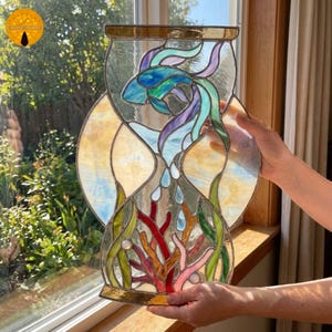 Könnte beinhalten: Ein Buntglasfenster in Sanduhrform mit einem blauen und violetten Kampffisch im oberen Bereich sowie roter und rosa Koralle mit grünem Seegras im unteren Bereich, eingefasst in eine Metallstruktur mit klaren Glaselementen.